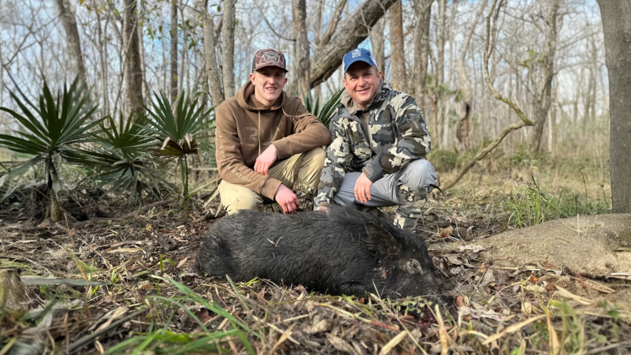 Two men crouch next to a large, dark-haired wild boar on forest floor. One wears camo, other brown. Bare trees and palmett...
