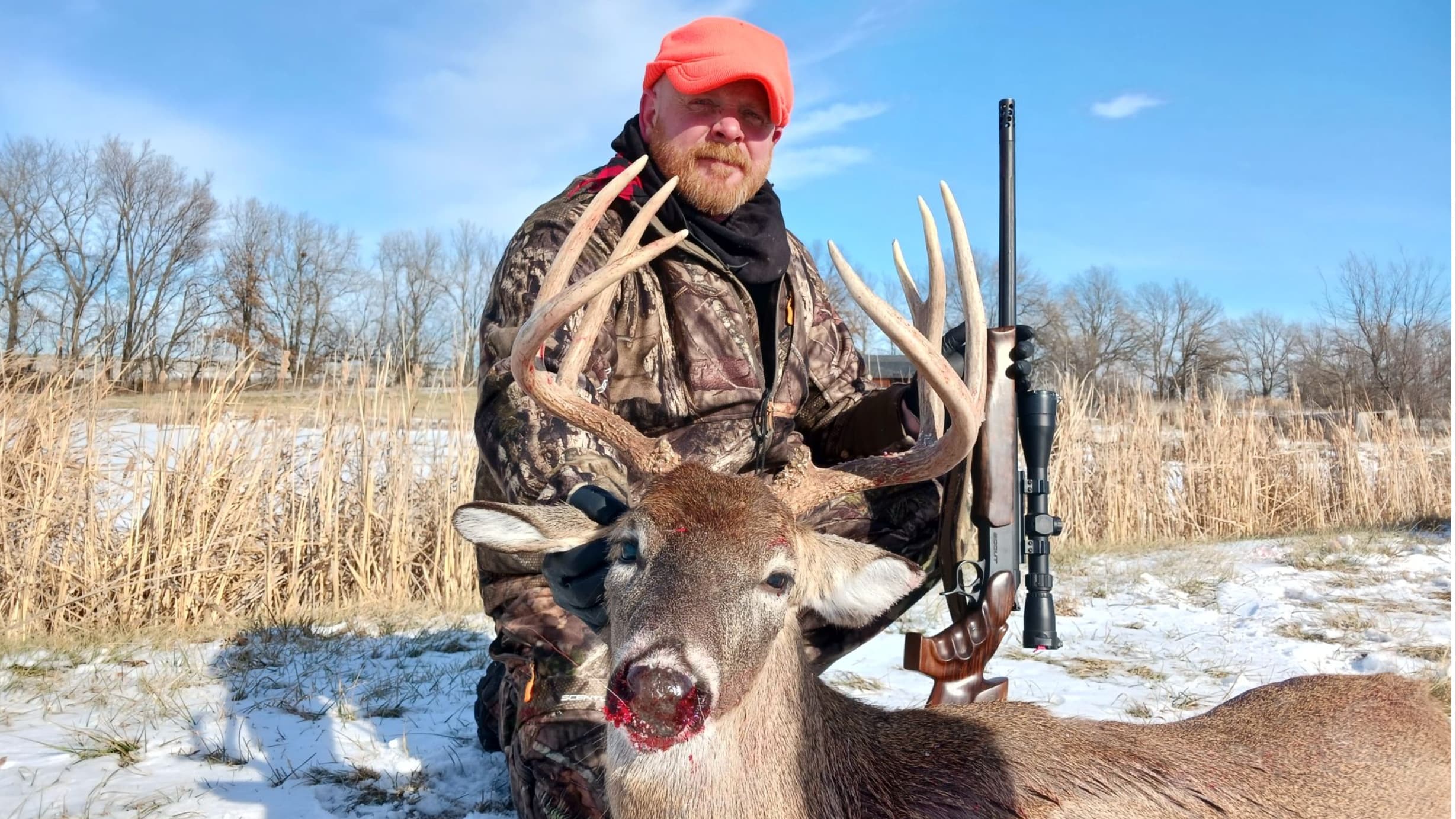 Hunter with deer. Man kneels beside a buck with large antlers, rifle on right. Winter scene, snow on ground, blue sky. Hun...