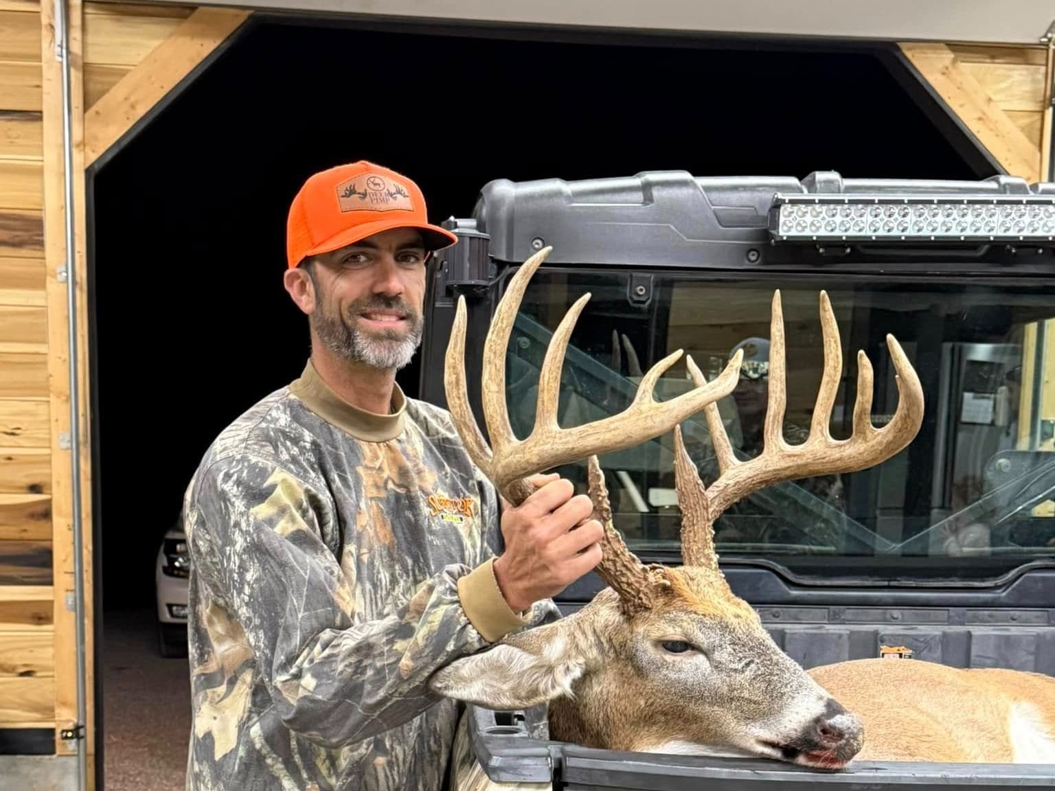 Hunter in camouflage and orange hat holds large buck antlers next to deer in a utility vehicle.