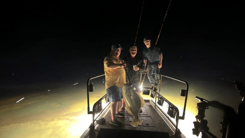 Three men on a boat at night, beaming light on the water. Large flat fish held up. Men smiling. Brightly lit water and dar...