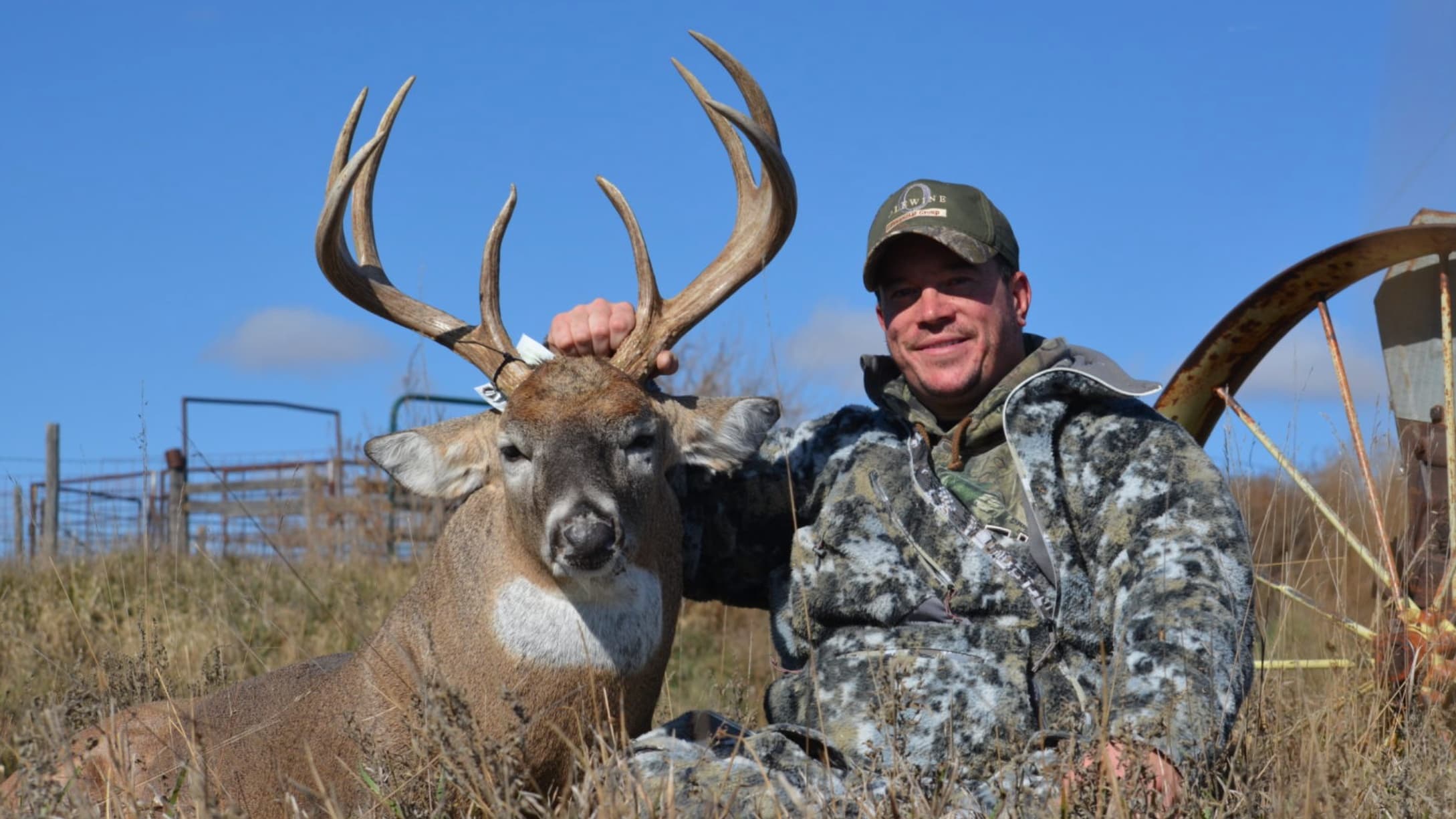 Man in camo holds large antlered deer. Bright blue sky backdrop. Man smiling, sitting in tall grass next to an old, rusty ...