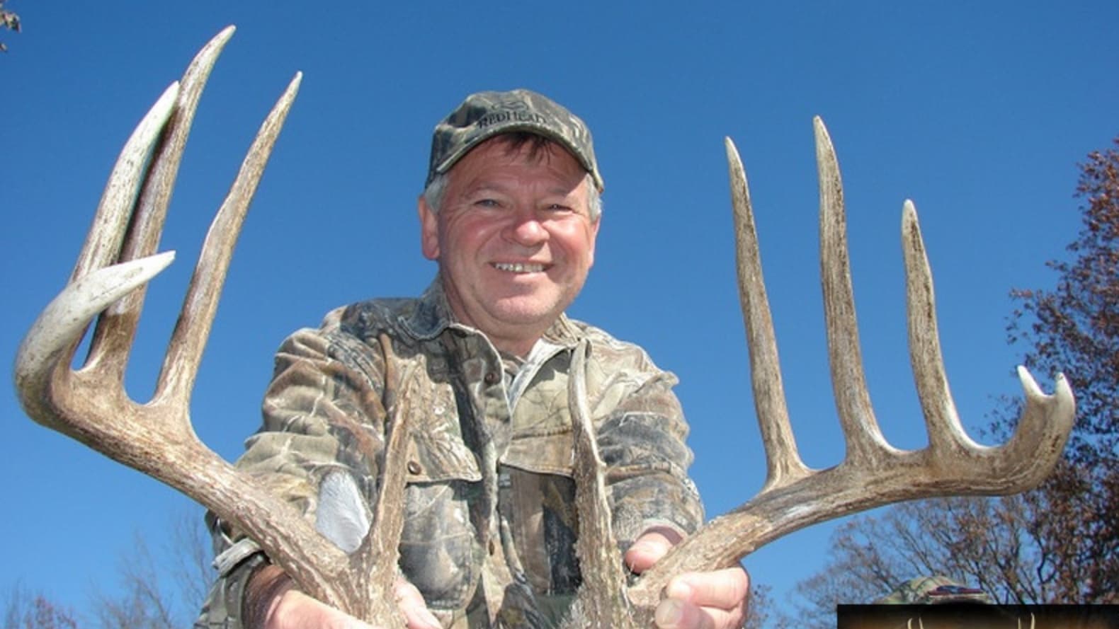Man in camouflage smiling, holding large deer antlers against a blue sky.