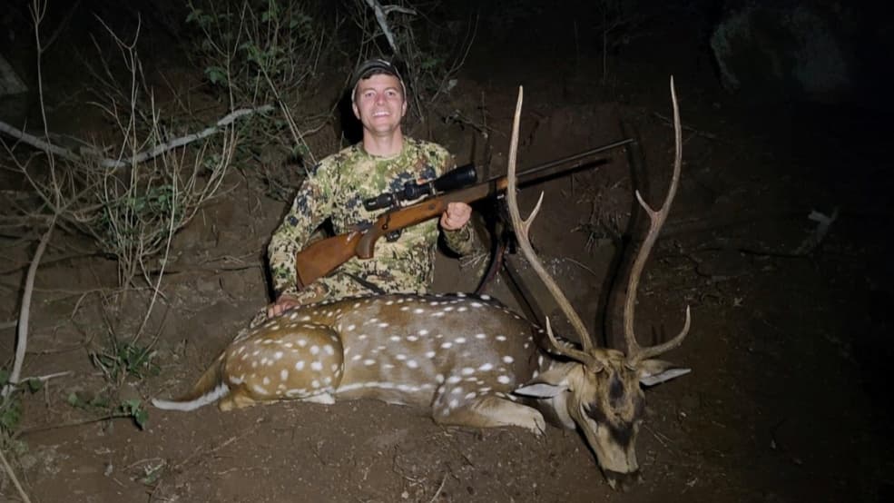 Hunter with rifle poses next to a spotted deer with large antlers. Man wears camo. Deer lies on ground. Dark background.