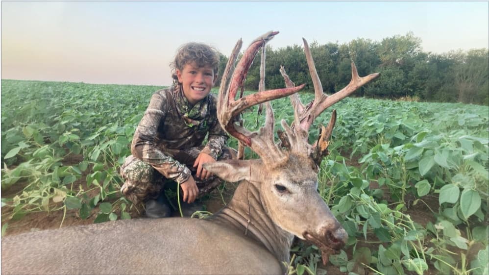 Smiling boy in camo kneels next to a large buck with impressive antlers. They are in a field of green plants.