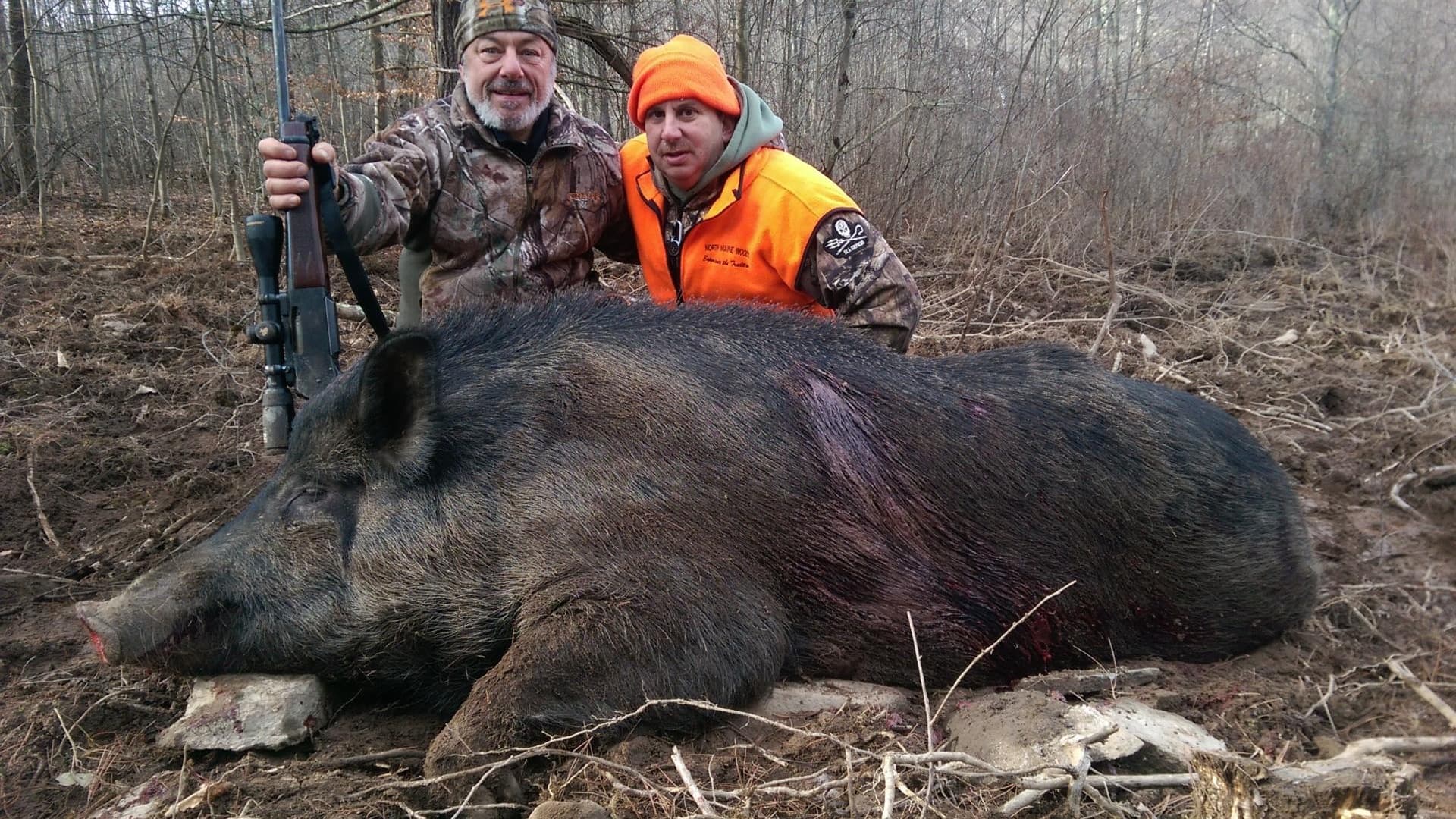 Two men pose with a large, dead wild boar. One man holds a rifle. The boar is dark with bristly hair, lying on muddy ground.