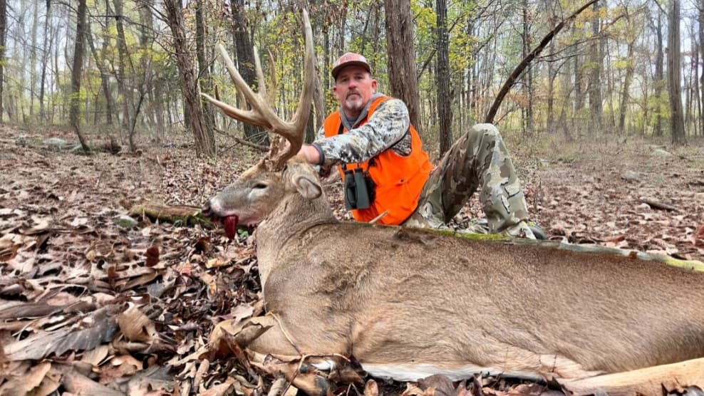 Hunter kneels beside a large buck with antlers in a forest. The buck lays on the ground, with blood visible. Hunter wears ...