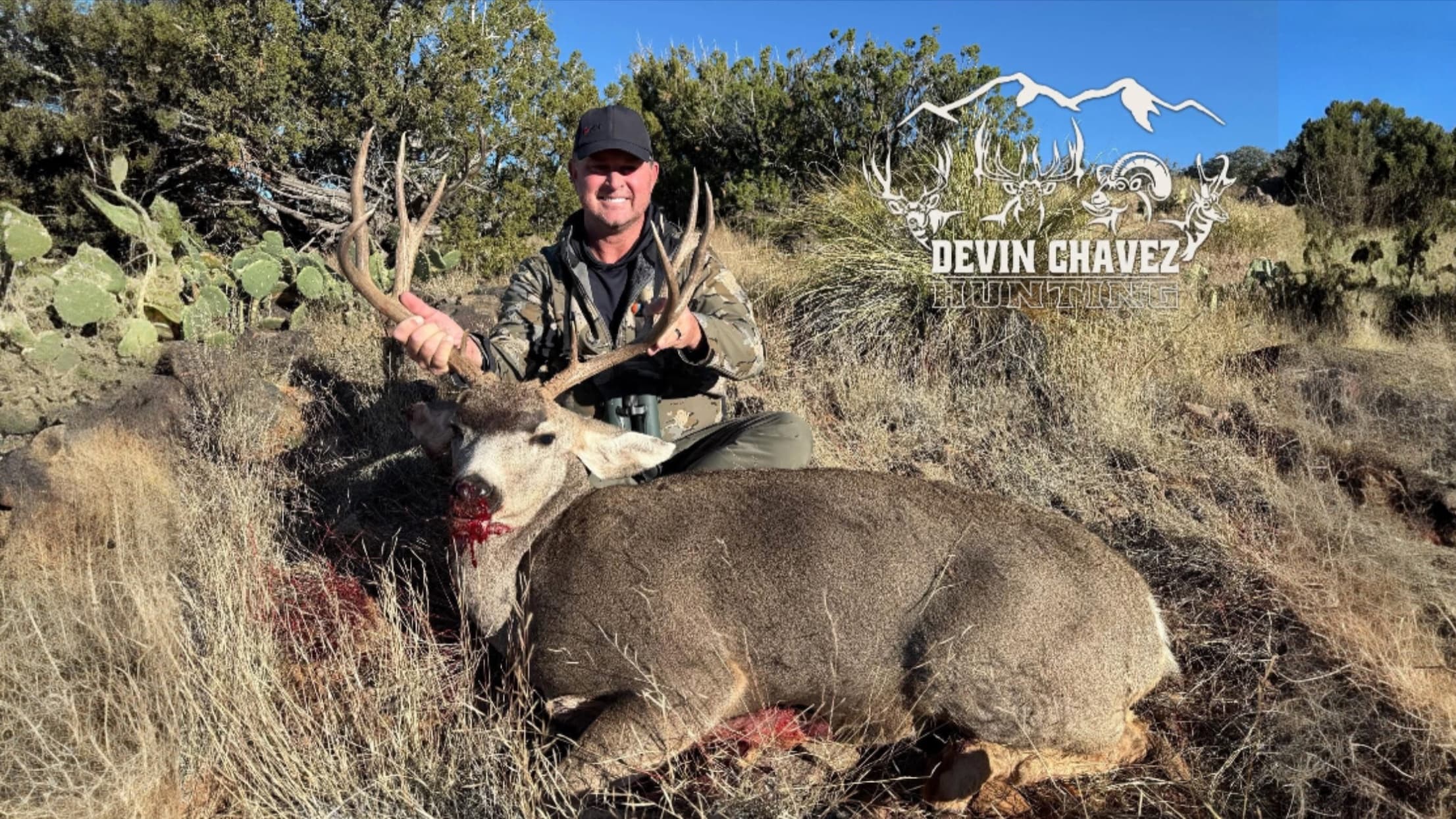 Man in camouflage holds up antlers of a large buck lying on the ground, blood visible. Background is arid, with scrub brus...