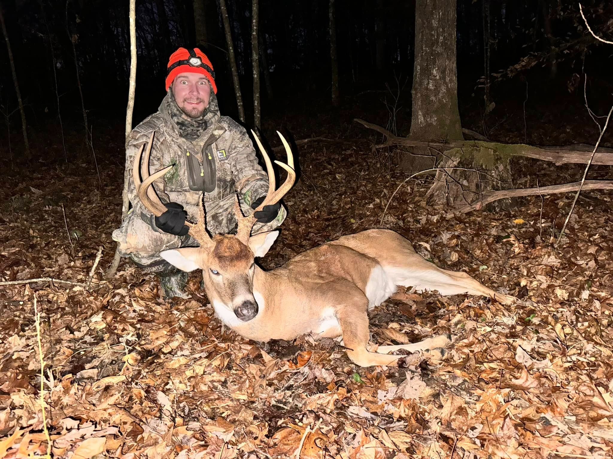 Hunter in camouflage and orange hat kneels by a fallen buck with large antlers on a forest floor covered in autumn leaves.
