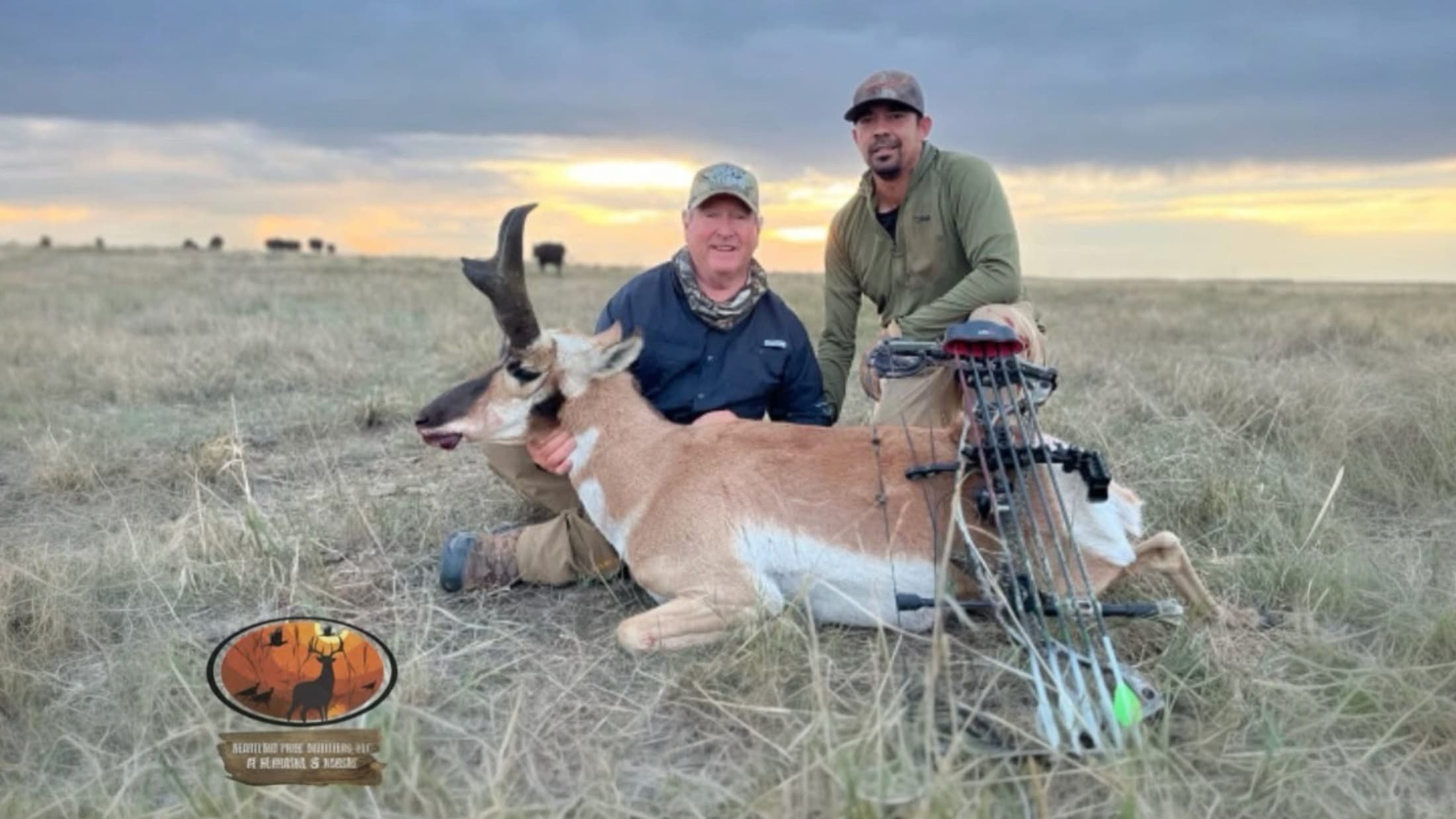 Two men kneel next to a deceased pronghorn. A bow rests beside the animal. Prairie grass and a cloudy sky are in the backg...